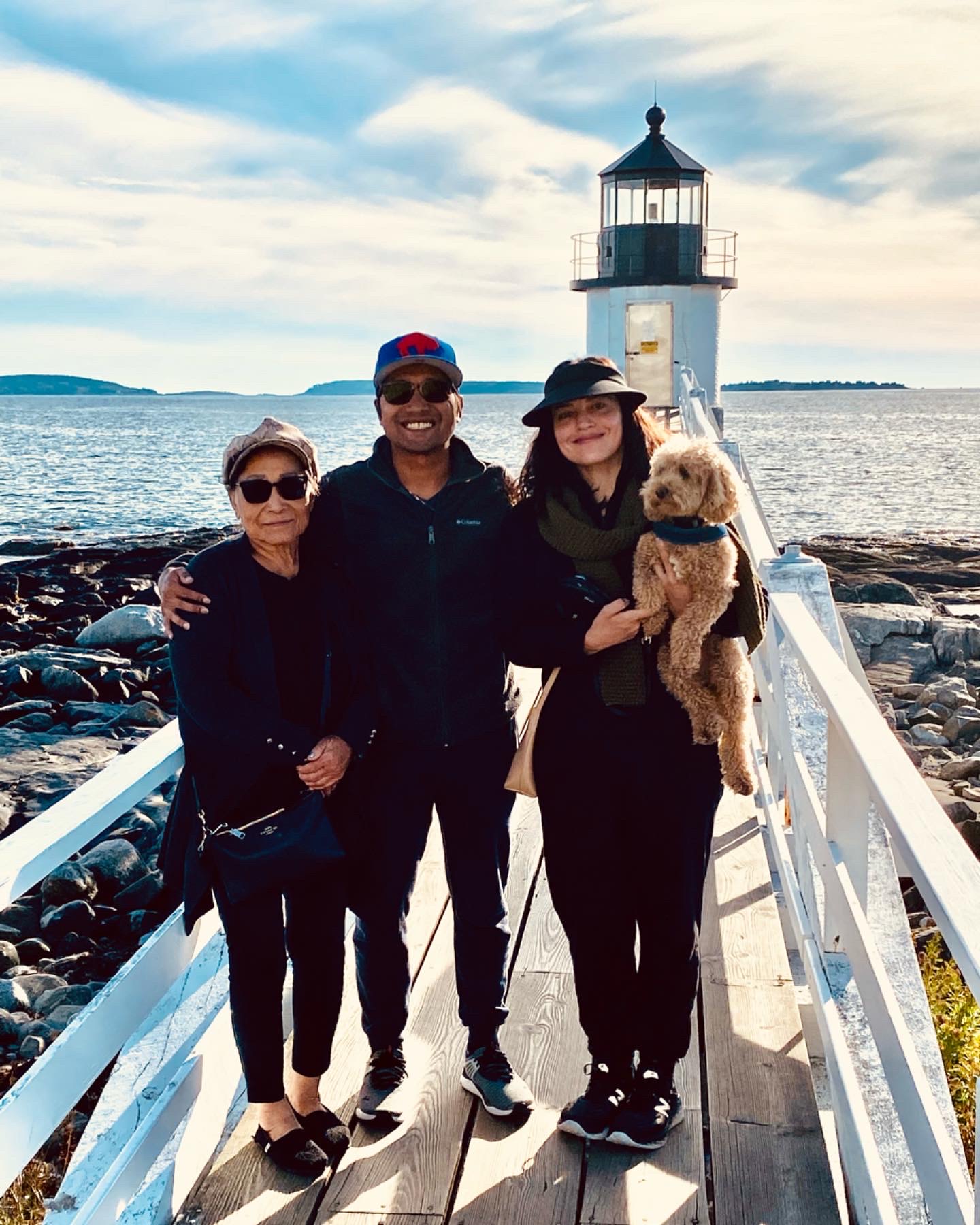 Three adults and a small dog posing on a walkway in front of a coastal lighthouse.