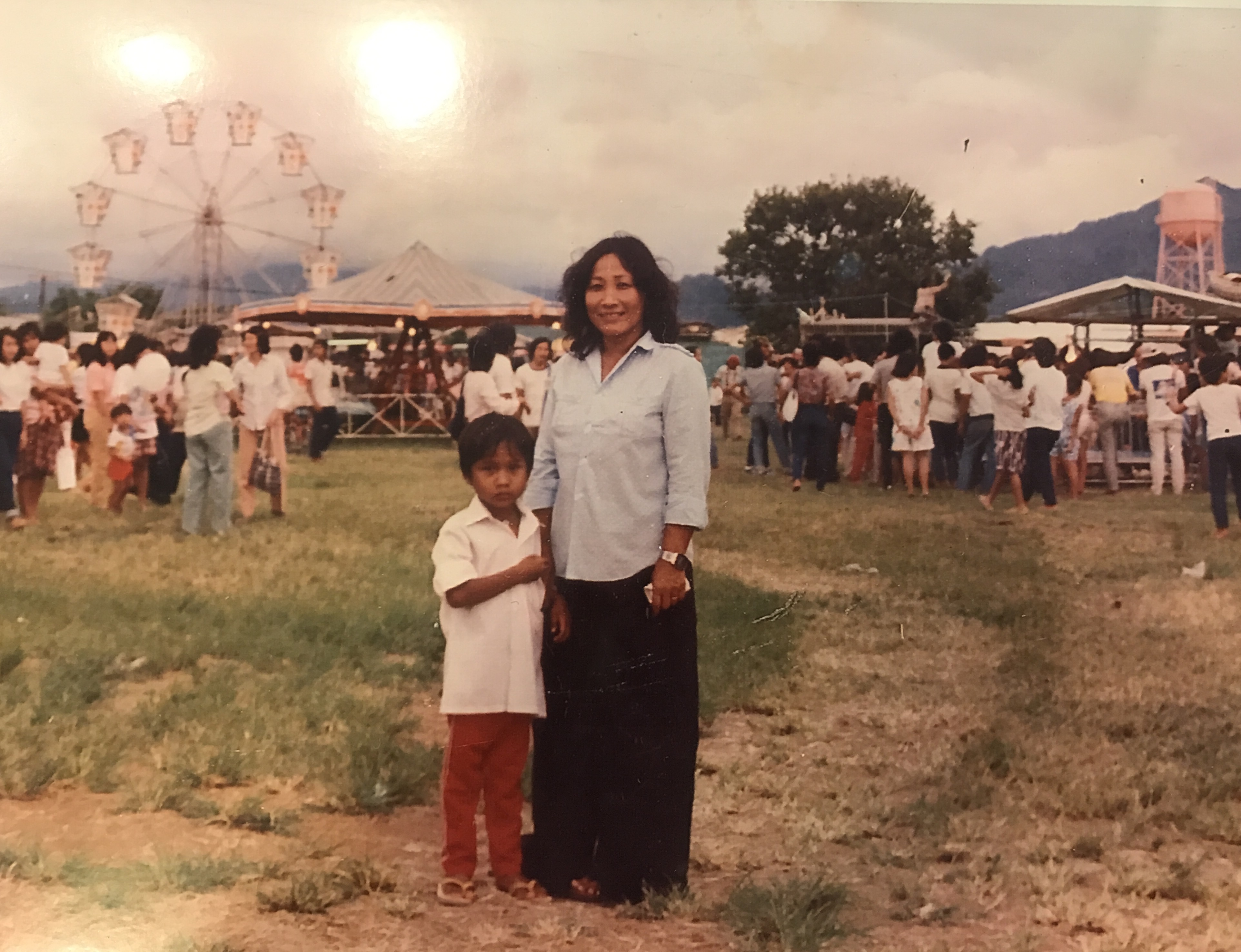 A young Minh standing beside his mother at a crowded outdoor fairground in the Philippines.