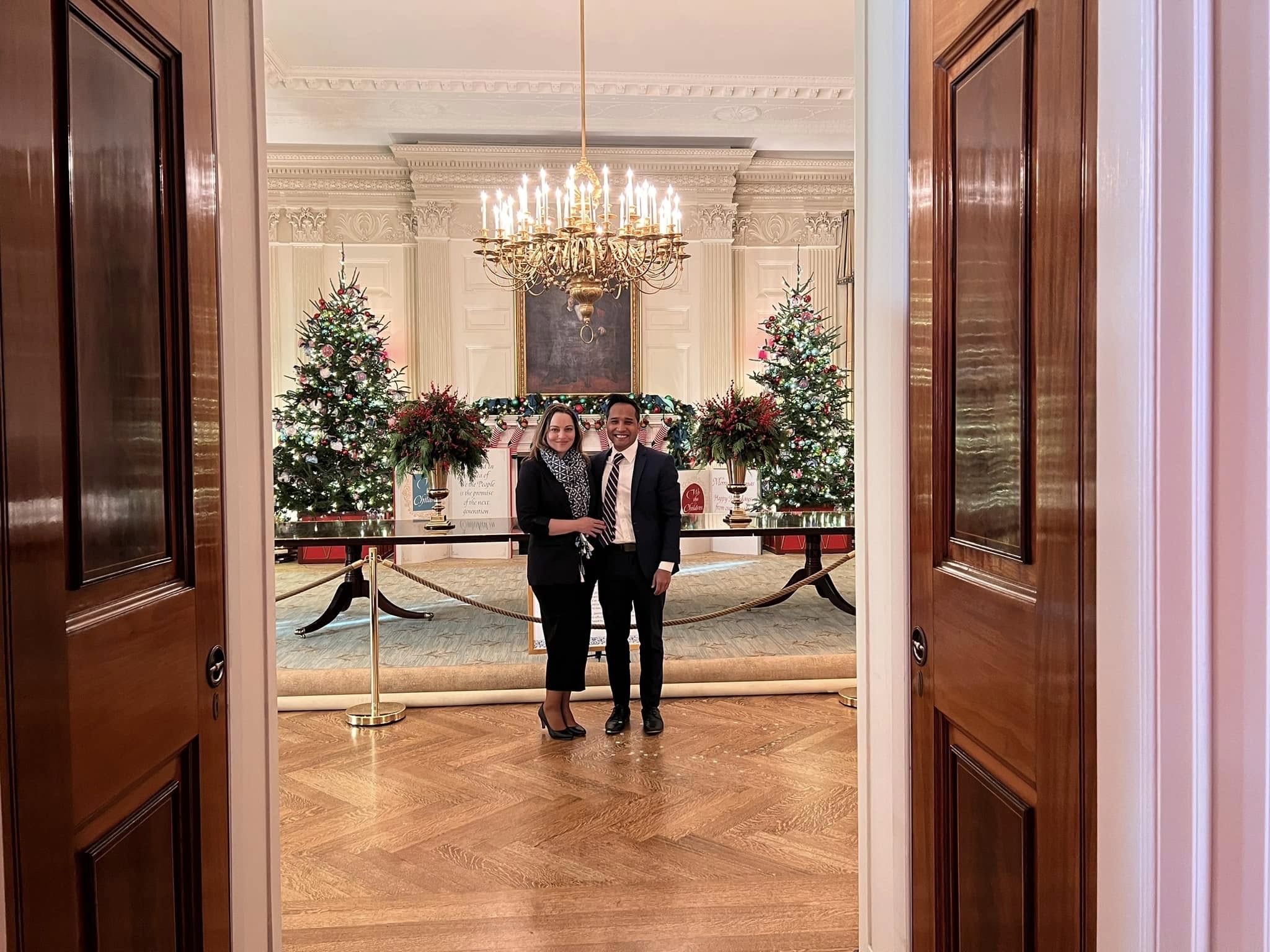 Two people standing together inside the White House during the holiday season with decorated Christmas trees and a chandelier behind them.