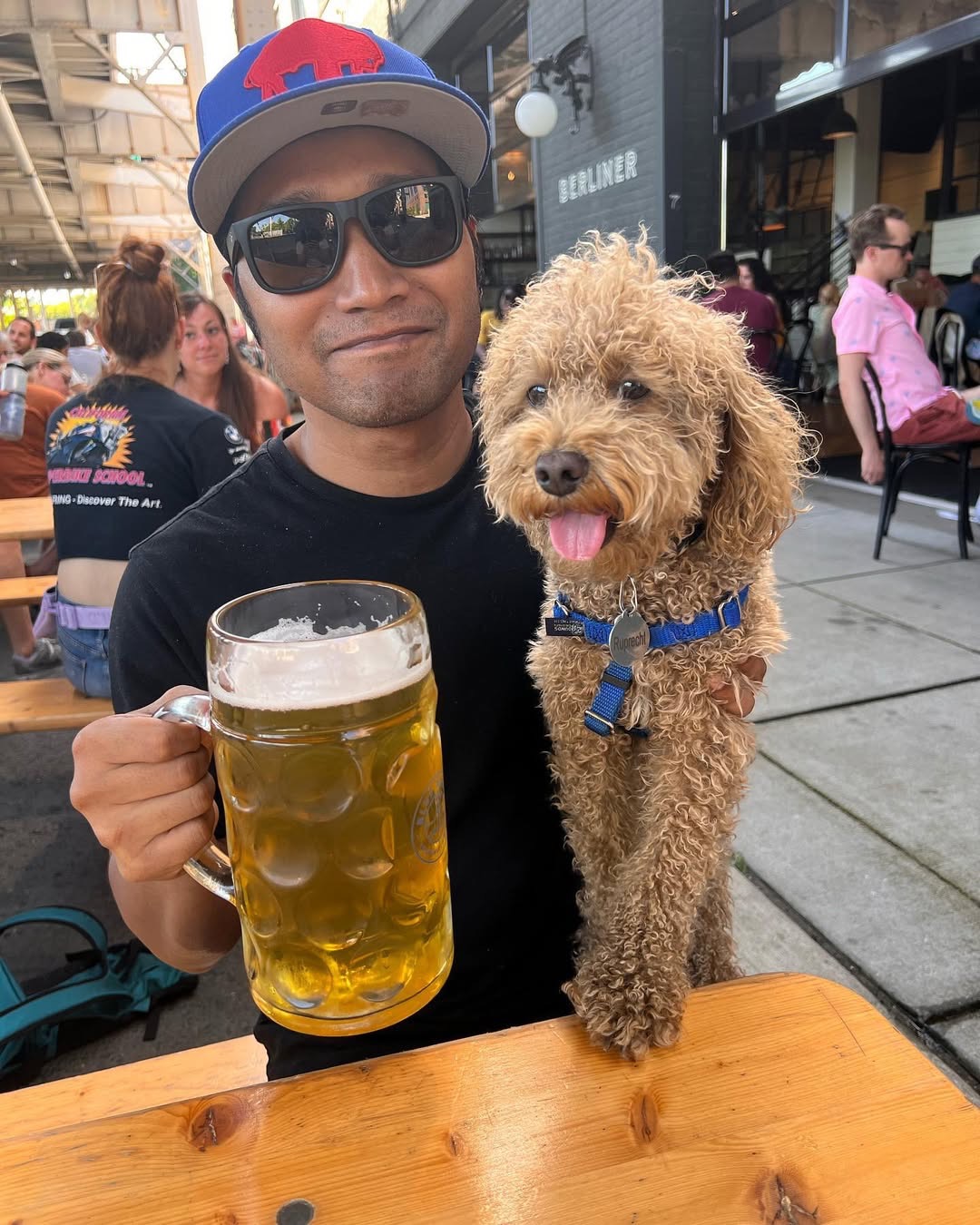 Man holding a beer mug while sitting with a small curly-haired dog at an outdoor table.