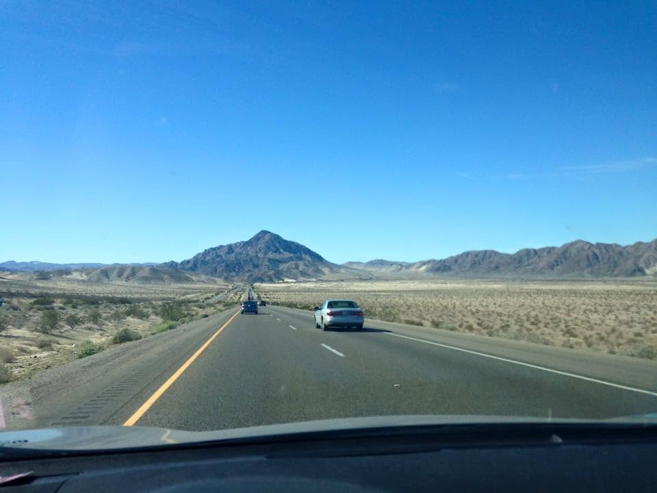 View from a car driving along a highway through a desert landscape toward distant mountains.