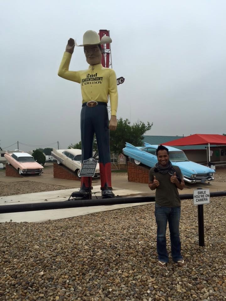 Person posing beside a giant cowboy roadside attraction and vintage cars.