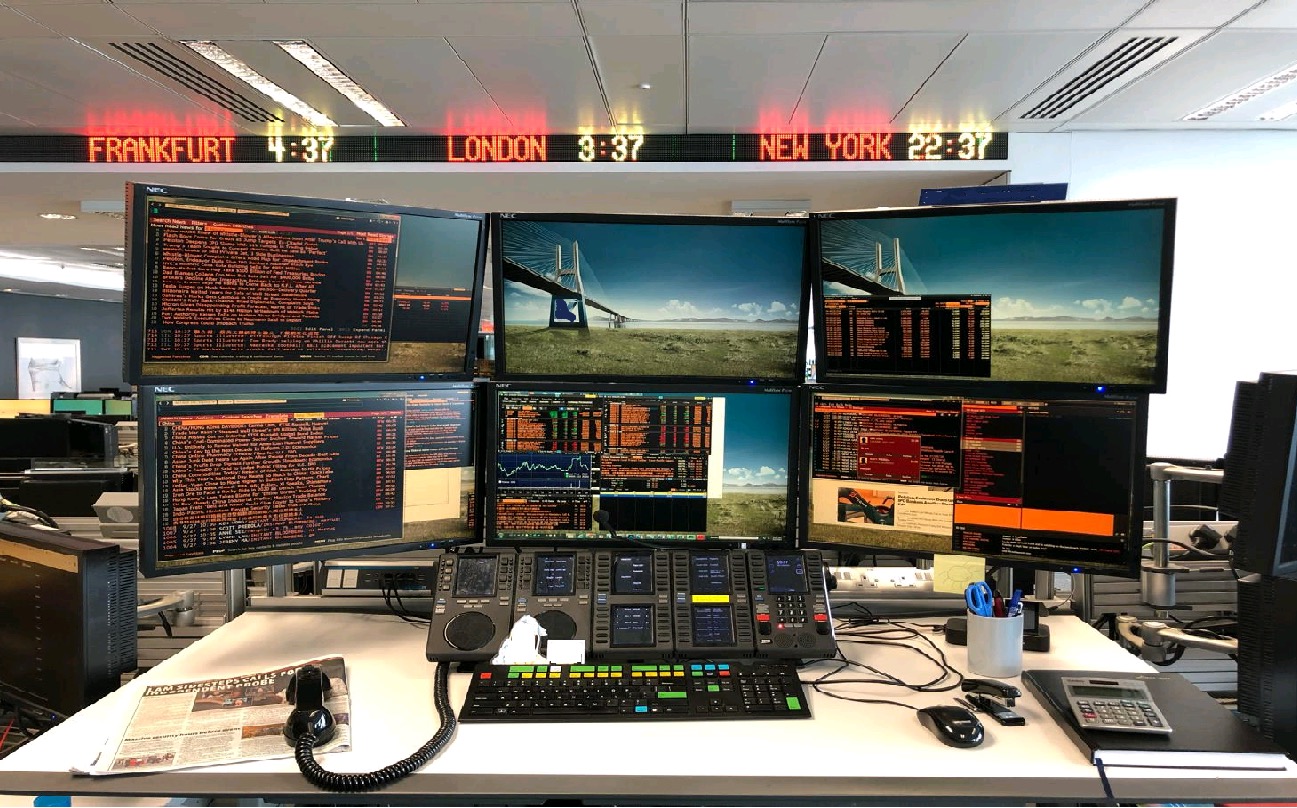 Financial trading workstation with multiple market-data monitors, keyboard, phones, and market clocks.