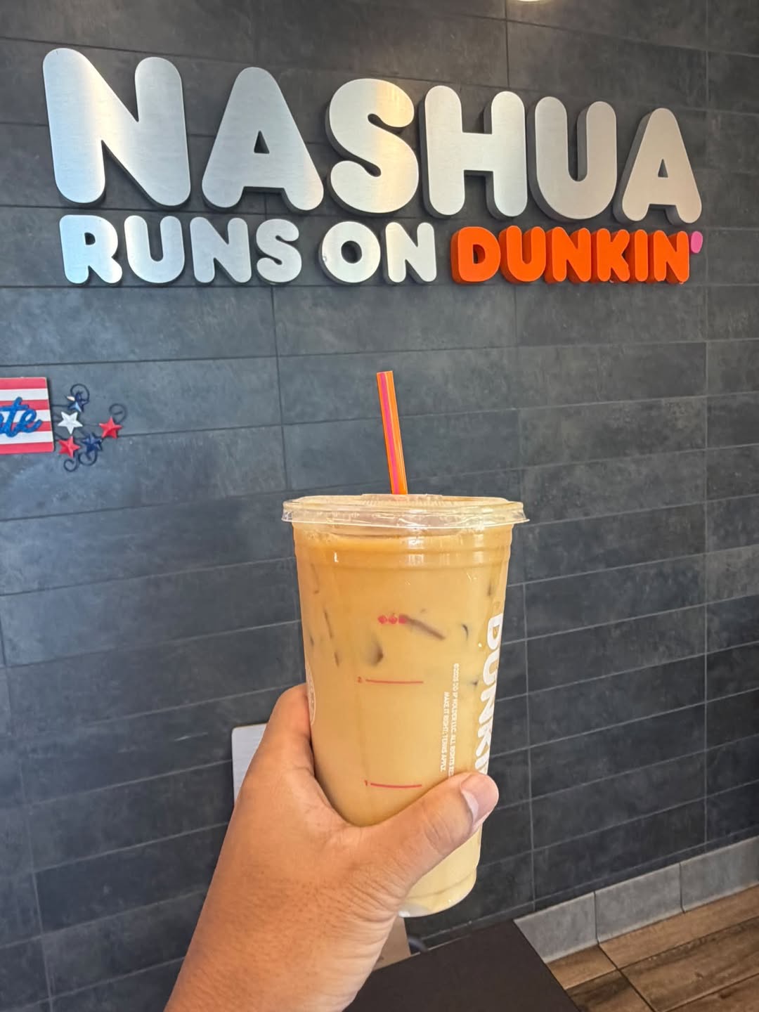Iced coffee held in front of a “Nashua Runs on Dunkin’” wall sign.