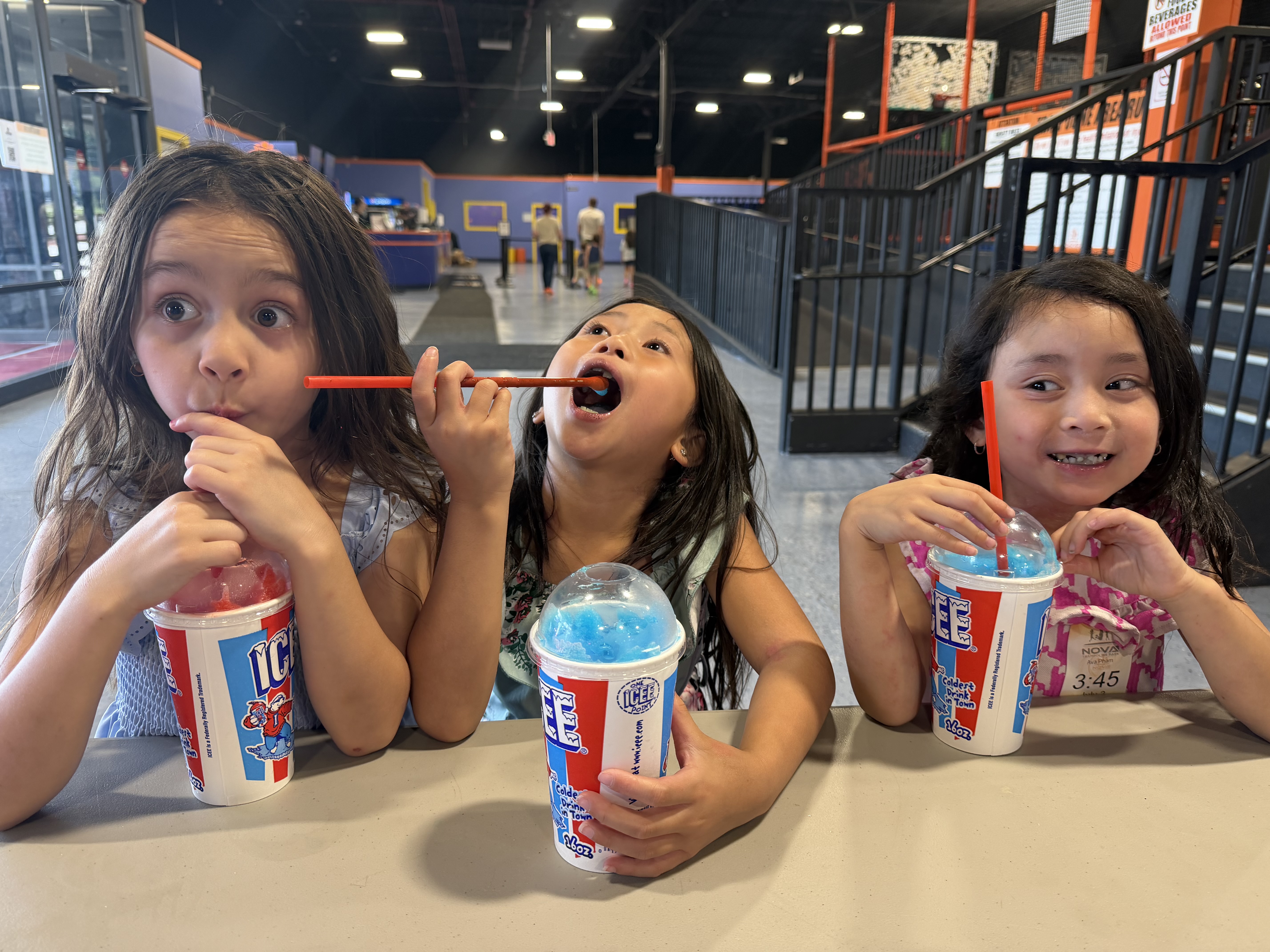 Three children making playful faces while enjoying colorful frozen drinks.