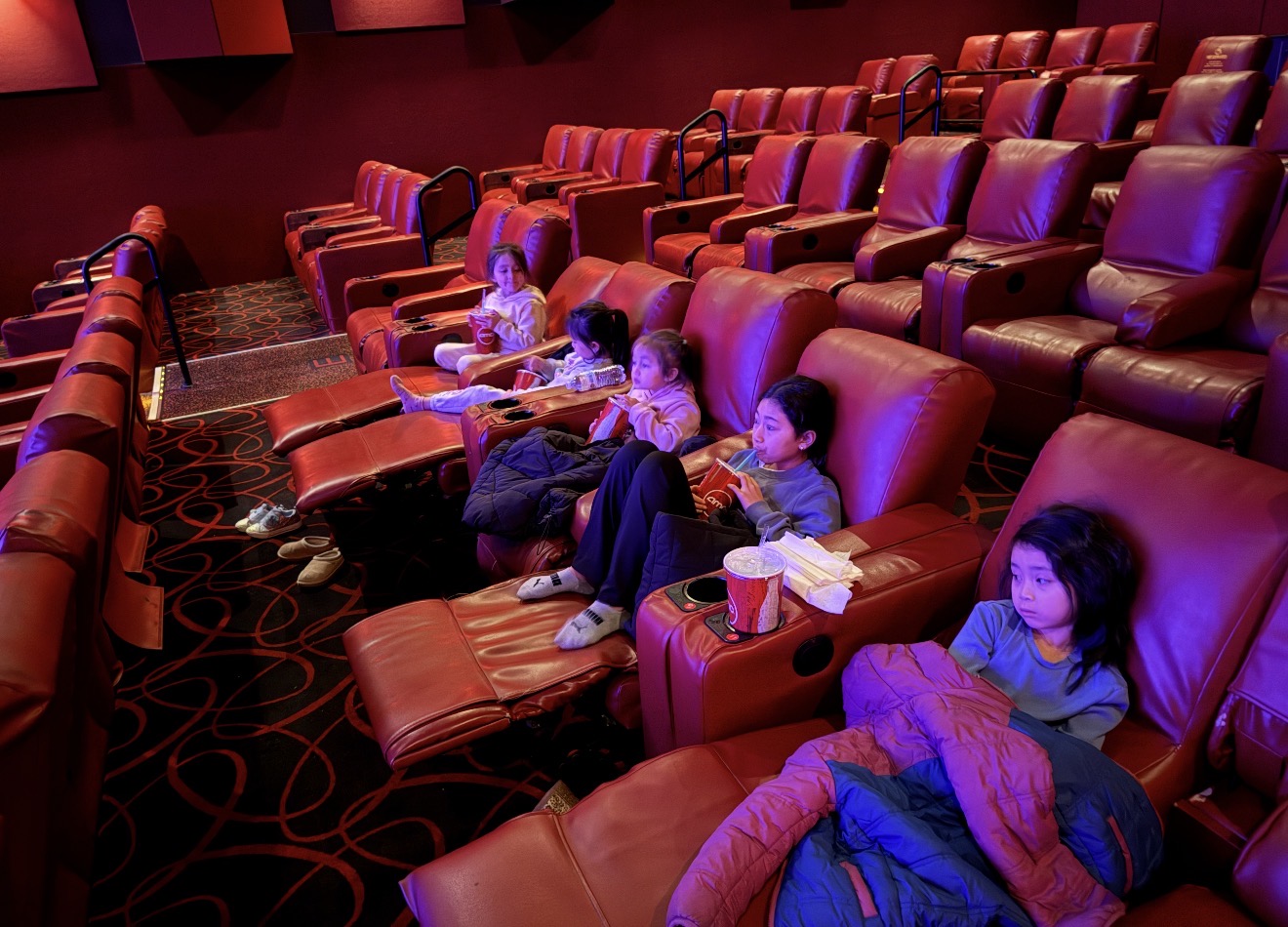 Five children relaxing in red recliner seats inside a movie theater.