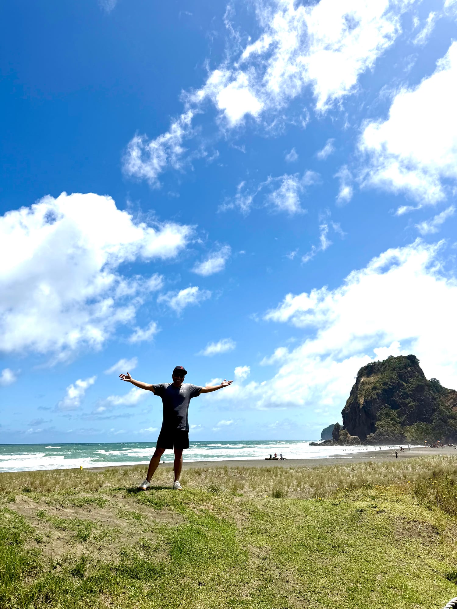 Person standing with arms out at Piha Beach under a blue sky.