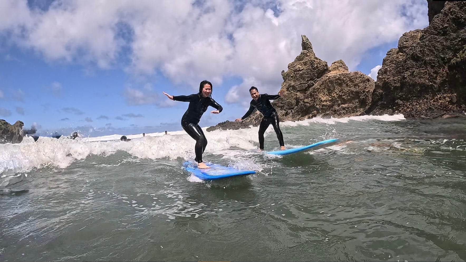 Two surfers riding small waves near rocky cliffs at Piha Beach.