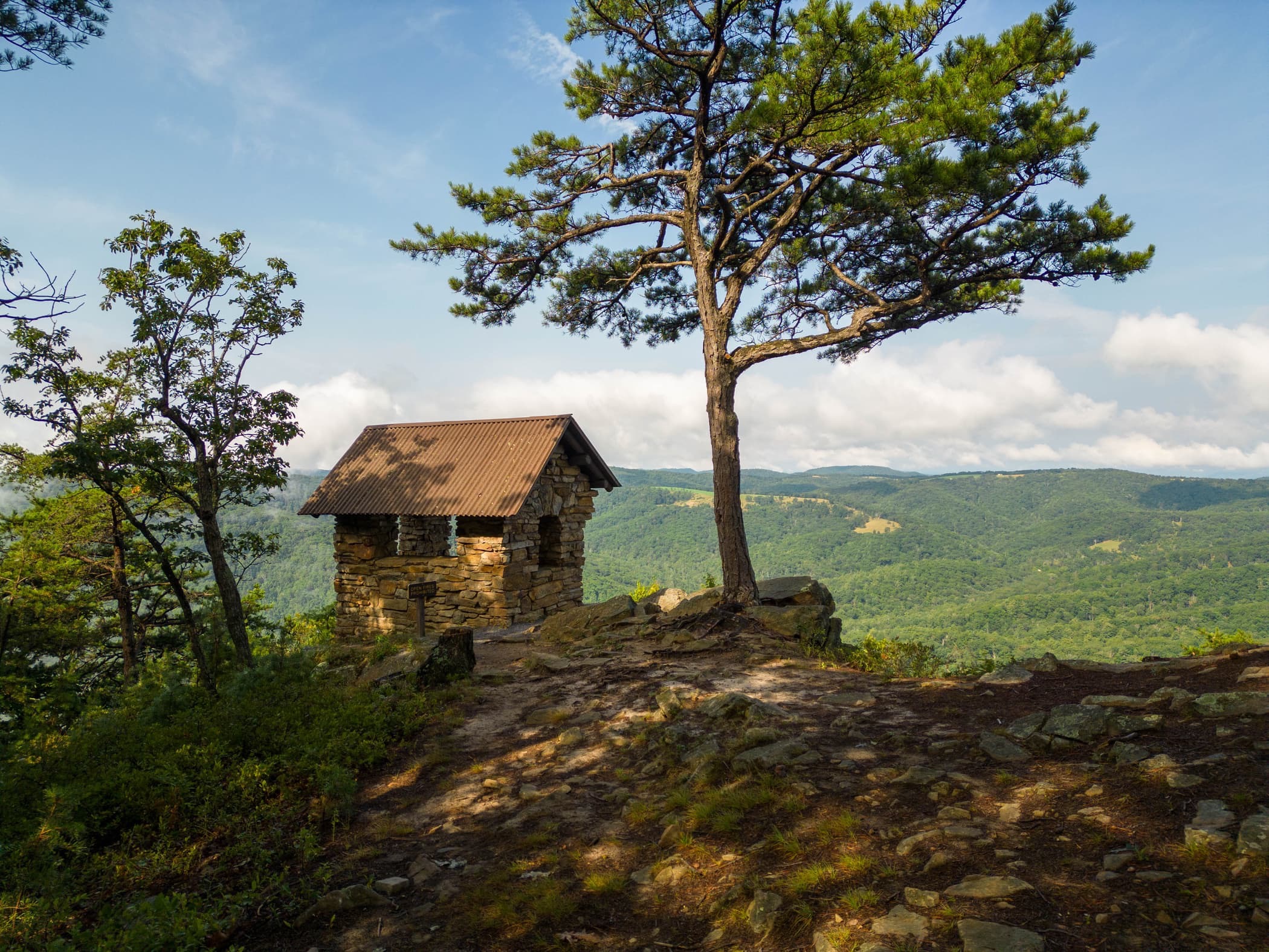 A stone overlook shelter beneath a pine tree with West Virginia mountains in the distance.